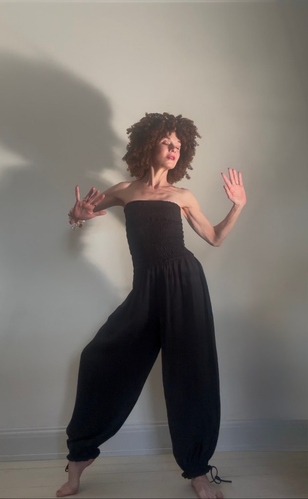 En kvinna med lockigt hår poserar i en svart jumpsuit mot en ljus vägg.
